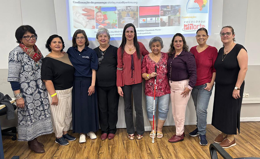 Nove mulheres estão juntas em um ambiente interno, posando para uma foto de grupo em frente a uma tela de apresentação. Uma mulher segura uma bengala branca. Elas estão sorrindo, vestidas com uma variedade de roupas casuais e profissionais.