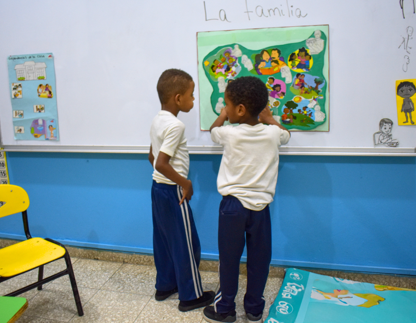 En una clase, dos chicos jóvenes examinan en una pizarra un colorido póster sobre la familia. La pared está decorada con dibujos y material educativo. A la izquierda se ve una silla amarilla.