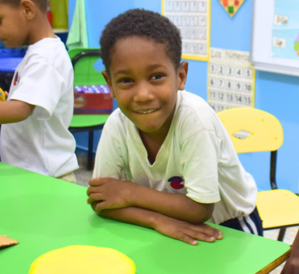 Wander sonríe apoyado en una mesa verde de una colorida clase. Lleva una camisa blanca y hay un proyecto de manualidades amarillo sobre la mesa frente a él. Al fondo se ven otros niños y material educativo.
