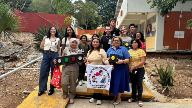 Los estudiantes del MIT D-lab y el personal del Centro de Atención Múltiple 1 están juntos fuera de la escuela. Algunas personas sostienen los dispositivos personalizados hechos para los estudiantes y una docente sostiene un cartel que identifica la escuela.