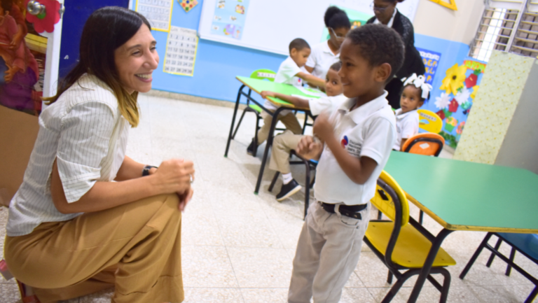 Una profesora sonriente se arrodilla junto a un niño en un aula mientras interactúan. Otros niños están sentados en mesas al fondo, participando en actividades. El aula es luminosa y colorida, con carteles educativos en las paredes.