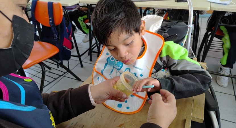 Un niño con cabello castaño oscuro, que usa un babero con un patrón azul, está sentado en una silla de ruedas en un salón de clases. Un adulto, que usa una mascarilla negra y tiene las manos visibles, sostiene una taza de plástico amarilla con una cuchara azul para que el niño coma de ella. El niño mira hacia abajo, hacia la taza, con la boca ligeramente abierta, en el acto de comer. El adulto sostiene la taza con ambas manos, mientras que el niño sostiene la cuchara azul en su mano derecha, llevándola a la taza. El fondo está desenfocado, mostrando sillas, escritorios y otros estudiantes. La iluminación es natural.
