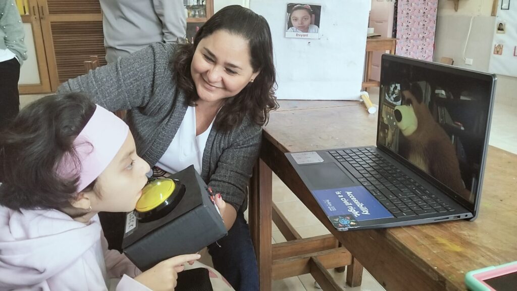 Una mujer sonriente observa a una niña que utiliza un dispositivo de ayuda con un gran botón amarillo e interactúa con un ordenador que muestra un dibujo animado de un oso en la pantalla. Están en un aula.