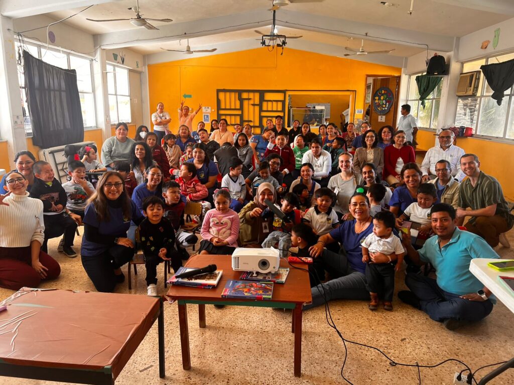 Un nutrido grupo de adultos y niños posan y sonríen para una foto de grupo en el interior de una sala muy iluminada con paredes amarillas, ventanas y mesas con libros y un proyector en primer plano.