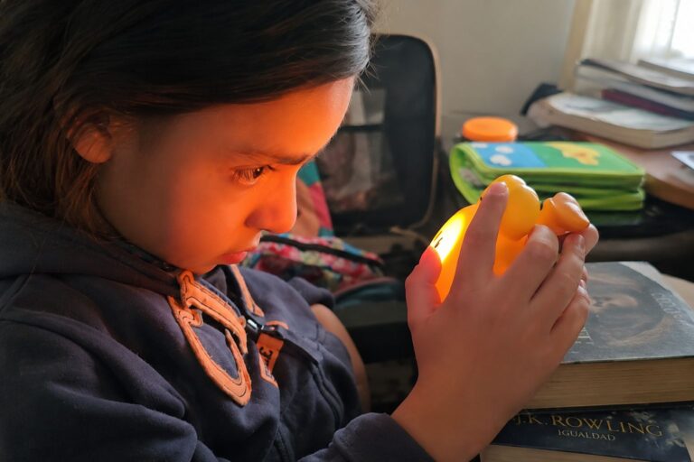 Un niño examina atentamente un patito de goma amarillo brillante mientras está sentado junto a una pila de libros. La habitación está suavemente iluminada y al fondo se ve material escolar.