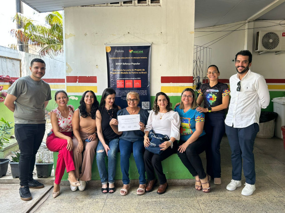 Um grupo de dez adultos sorridentes, nove mulheres e um homem, posam para uma foto ao ar livre em frente a uma entrada de prédio com um banner colorido. A maioria está sentada em um banco baixo verde, e dois estão de pé. No centro, uma mulher segura um documento de certificado. O banner atrás deles inclui os logotipos da Perkins e do município de Macapá. A atmosfera é comemorativa e de orgulho.
