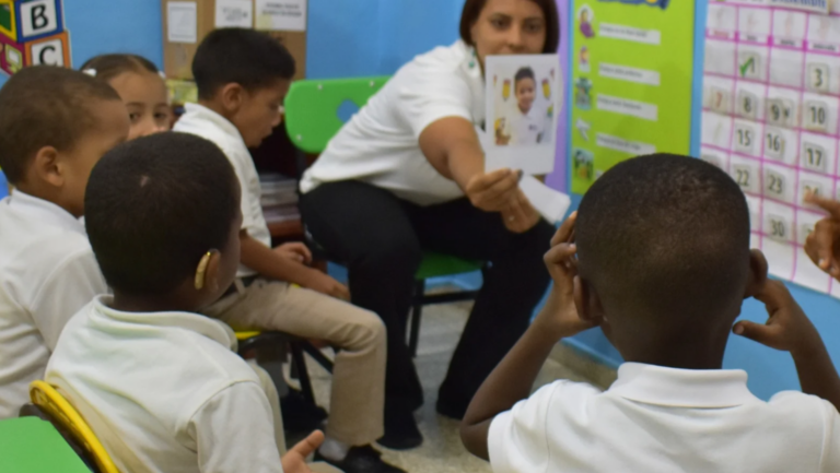 Una docente se sienta con alumnos jóvenes en un aula, sosteniendo una tarjeta. Los niños y niñas, todos con camisas blancas, se sientan en semicírculo, prestando atención a la docente que está al frente.