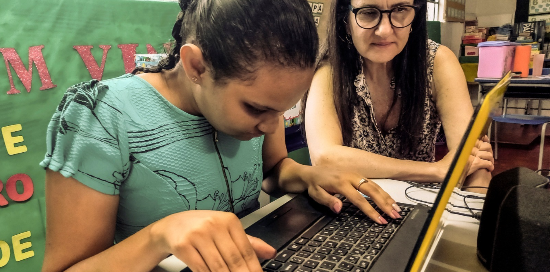 Una niña utiliza un ordenador portátil con las manos en el teclado, mientras una mujer se sienta a su lado, observando y apoyando. Están en un aula con coloridos adornos en la pared.