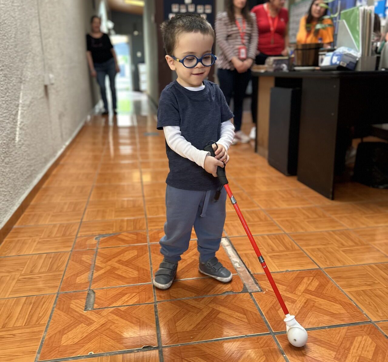 Un joven con gafas utiliza un bastón blanco para desplazarse por un pasillo de baldosas marrones. Varios adultos le observan desde el fondo, cerca de un escritorio.