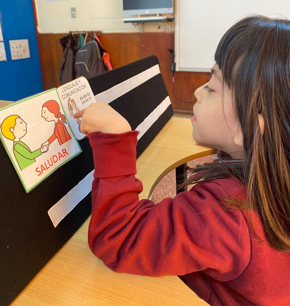 Una joven en un aula señala una tarjeta con texto, sostenida sobre una pizarra que muestra una ilustración de dos personas dándose la mano con la inscripción "SALUDAR". Al fondo, una pizarra y una computadora.