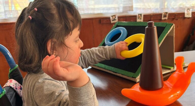 Una niña se sienta en una mesa, coge unos anillos de colores y un juguete en forma de cono, participando en una actividad lúdica y educativa.