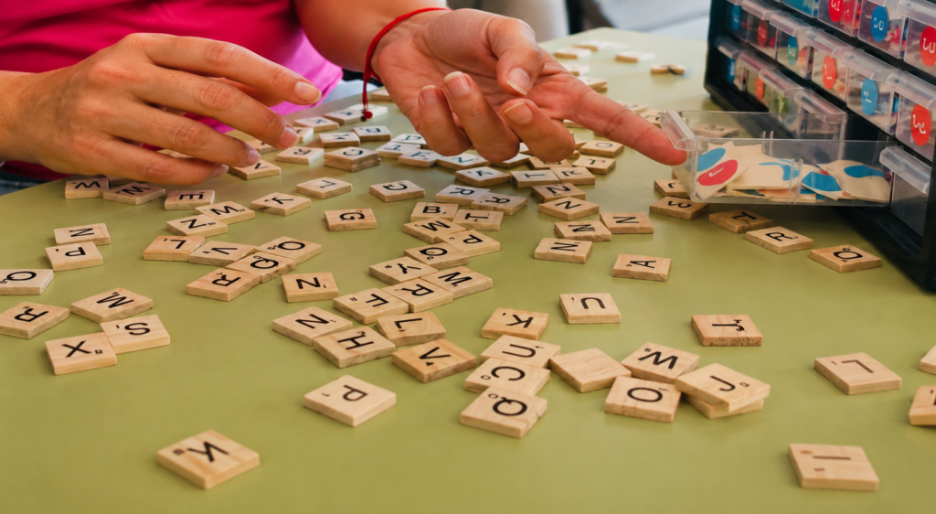 Una persona coloca fichas de letras de madera sobre una mesa verde, con las manos extendidas hacia un cajón que contiene fichas de juego rojas y azules. Algunas fichas están esparcidas al azar por la superficie.