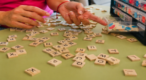 Una persona coloca fichas de letras de madera sobre una mesa verde, con las manos extendidas hacia un cajón que contiene fichas de juego rojas y azules. Algunas fichas están esparcidas al azar por la superficie.