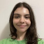 Uma mulher com longos cabelos castanhos e uma camisa verde sorri para a câmera enquanto está em frente a uma parede branca lisa.