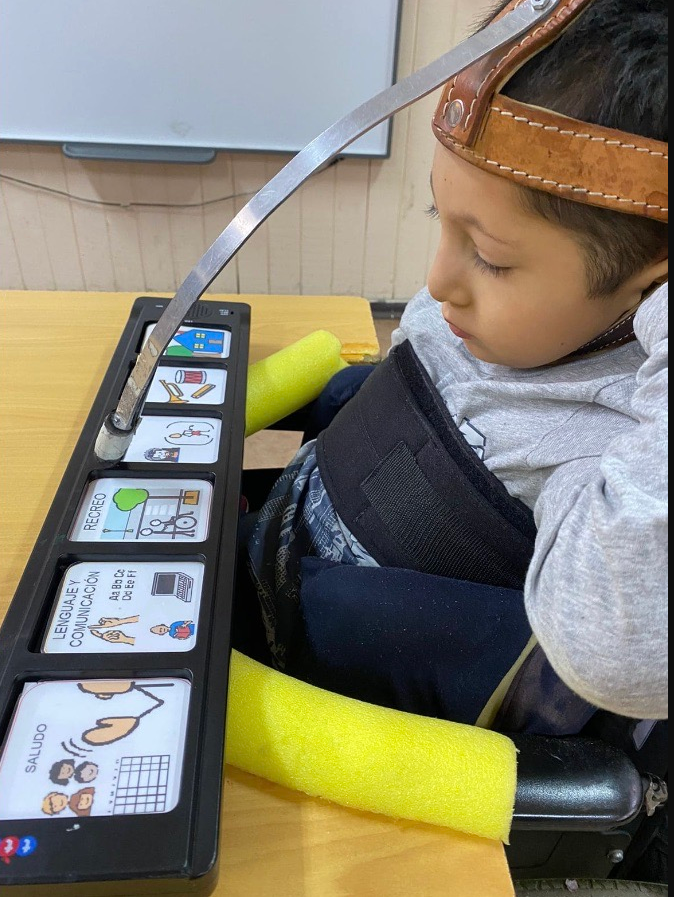 Un niño utiliza un puntero en la cabeza para interactuar con un tablero de comunicación que tiene tarjetas con símbolos de frases comunes. Está sentado en una silla de ruedas frente a un escritorio, con equipo de apoyo y reposabrazos de espuma amarilla.