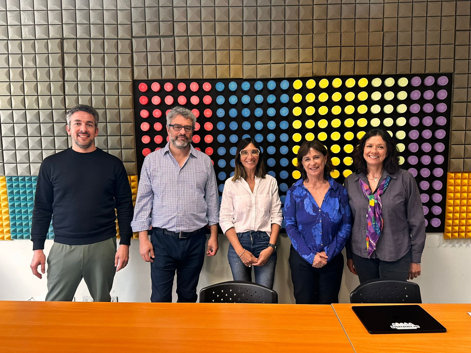 Cinco personas sonríen delante de un colorido mural con una cuadrícula de grandes puntos rojos, amarillos, azules y negros, detrás de una mesa de conferencias de madera en una sala moderna.