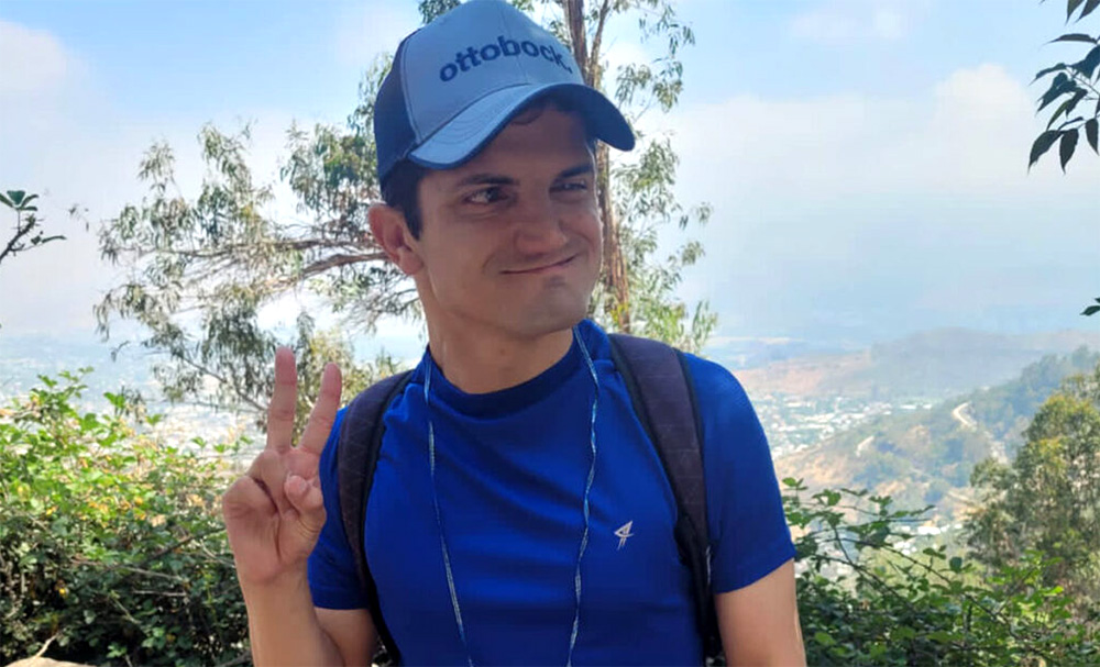 Un adolescente con camisa azul y gorra está de pie al aire libre, sonriendo y haciendo el signo de la paz con la mano. Lleva una mochila y una etiqueta con su nombre, con árboles y un paisaje montañoso de fondo.
