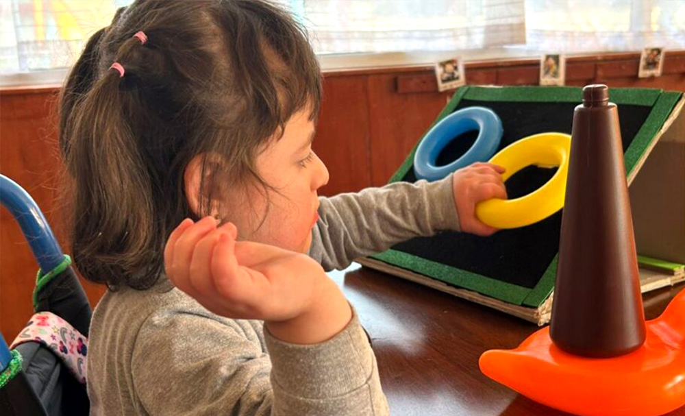Una niña se sienta en una mesa, coge unos anillos de colores y un juguete en forma de cono, participando en una actividad lúdica y educativa.