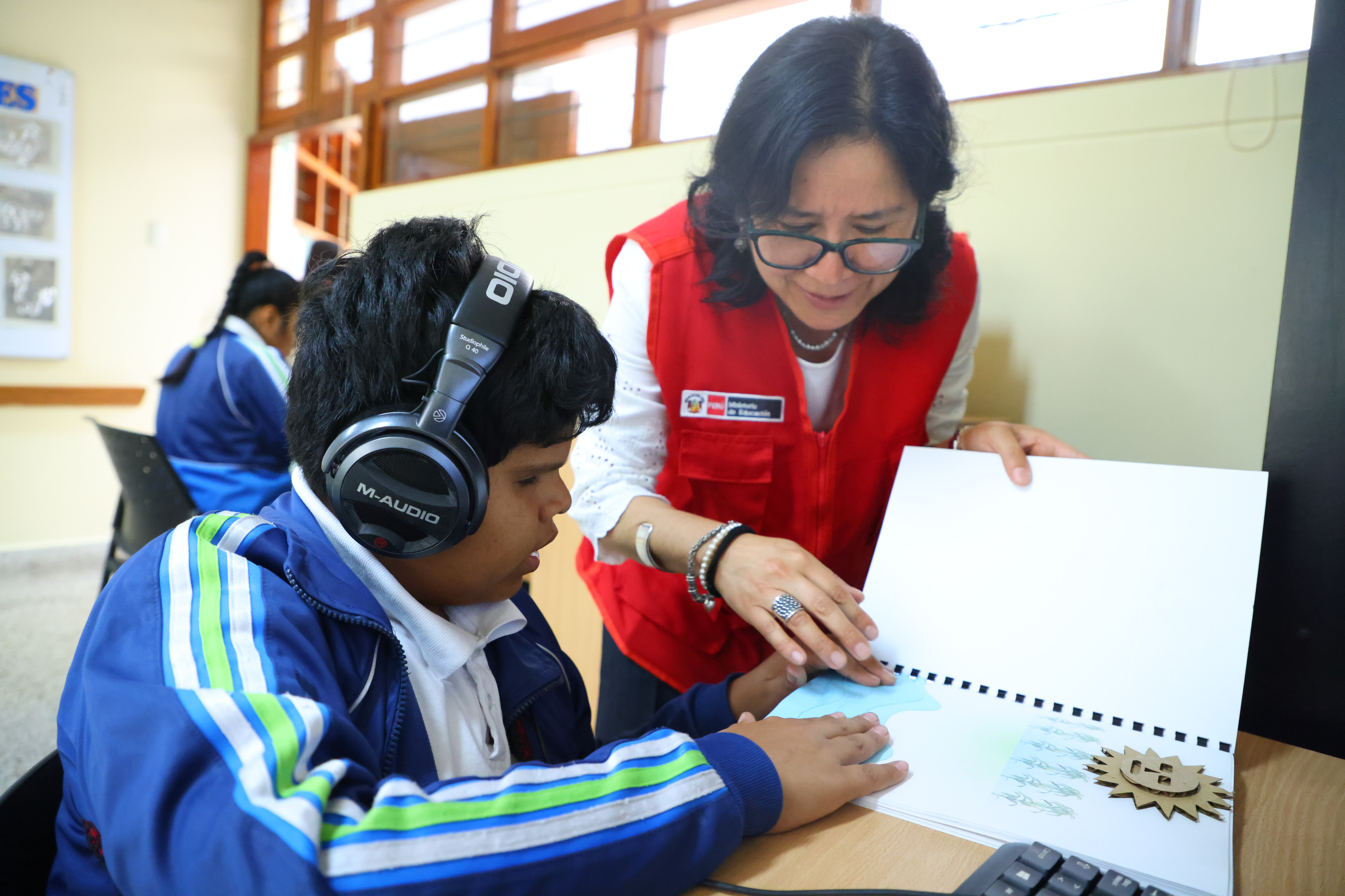 Una mujer con chaleco rojo ayuda a un niño con auriculares a tocar un gráfico táctil en un libro para alumnos con discapacidad visual en un aula.