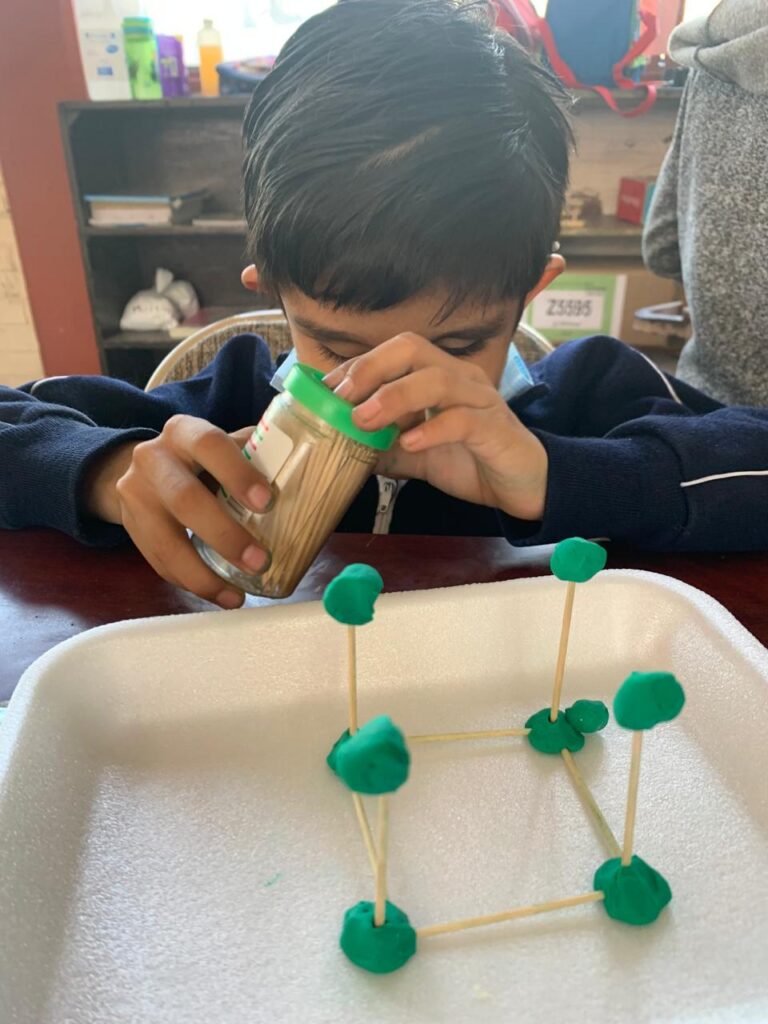 Christopher va sacando con cuidado palillos de un recipiente y construye una estructura tridimensional en forma de cubo con plastilina verde y palillos sobre una bandeja blanca.