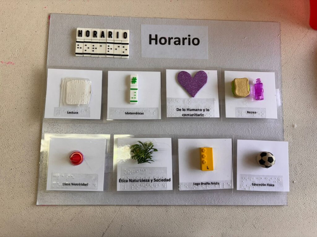 Un panel táctil con el horario escolar, con símbolos en relieve y braille para cada asignatura, que incluye objetos como un ladrillo de LEGO, un botón, un balón de fútbol de juguete y figuras con diferentes texturas, etiquetados en español con texto impreso y en braille.