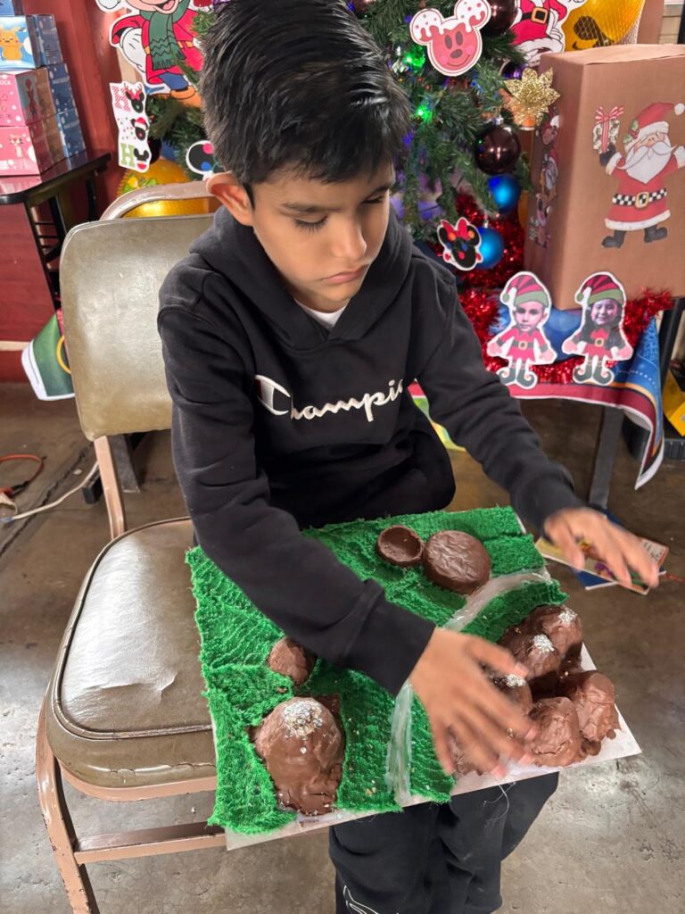 Un niño pequeño, vestido con una sudadera con capucha negra de la marca Champion, está sentado en una silla, haciendo una manualidad con piedras de arcilla marrones sobre una superficie verde. Detrás de él hay adornos navideños, entre los que se incluyen un árbol de Navidad y recortes festivos.