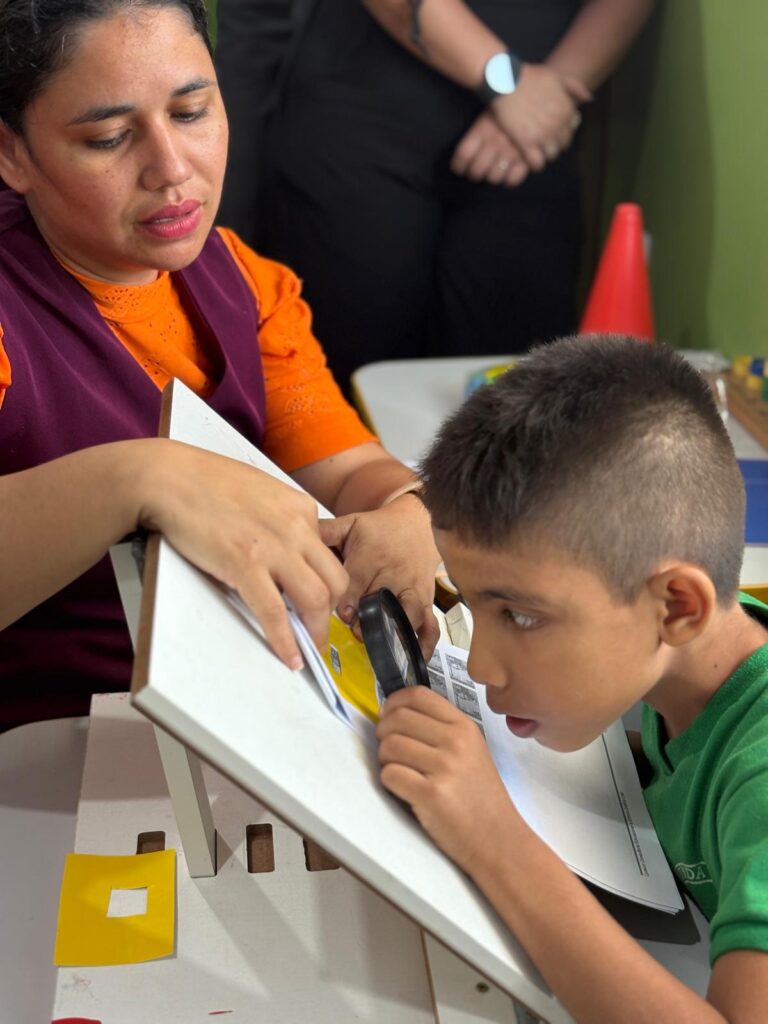 Efraim fazendo uma avaliação com uma professora capacitada pela Perkins. Ele utiliza um plano inclinado, uma lupa e um tiposcópio, auxílios fundamentais para tornar este teste acessível de acordo com suas necessidades educacionais.