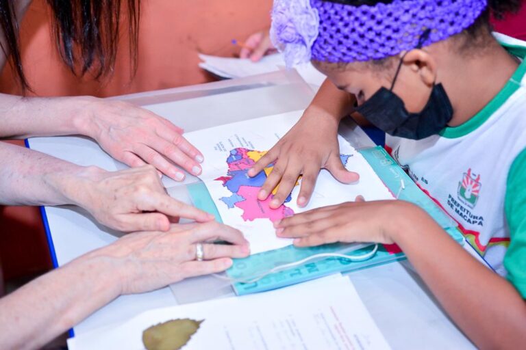 A foto mostra as mãos de uma estudante e de duas educadoras trabalhando juntas sobre uma mesa. À direita, a aluna usa uma faixa de crochê roxa no cabelo com uma flor, uma máscara cirúrgica preta e um uniforme escolar branco e verde. Suas mãos estão espalmadas sobre um mapa tátil adaptado e colorido do Brasil, explorando suas texturas e divisões regionais. À esquerda, as mãos das educadoras tocam o mesmo mapa com delicadeza, orientando a exploração tátil da aluna.
