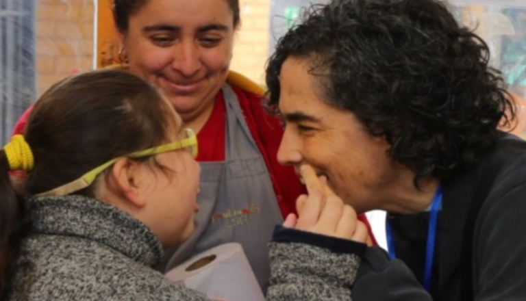 Una mujer y una niña con gafas, ambas sonriéndose el una al otro, comparten un momento de alegría. La niña está tocando la cara de la mujer cerca de su boca. Detrás de ellas, otra mujer las observa con una cálida sonrisa.