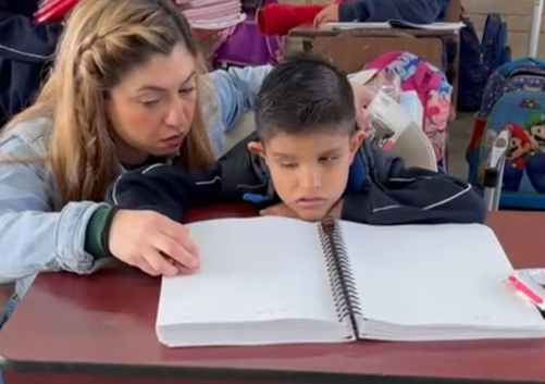 A woman sits beside a boy, guiding his hand as they read from a large Braille book on a desk in a classroom setting. The boy appears focused, and other children are visible in the background.