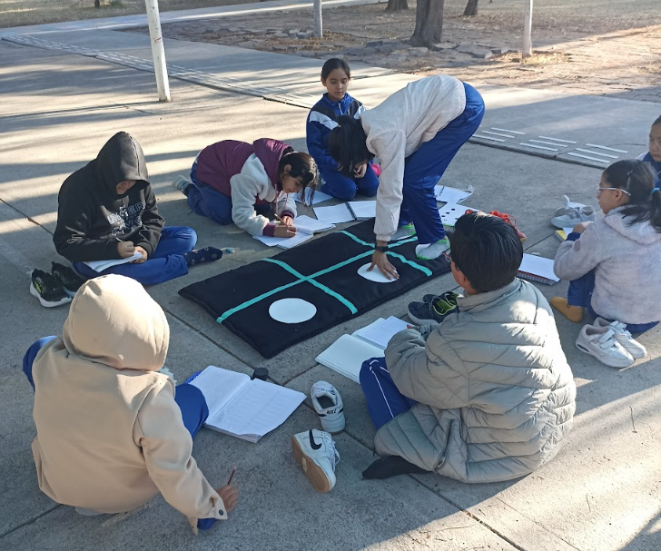 Un grupo de niños está sentado en el suelo al aire libre, reunido alrededor de un gran tablero. Algunos están escribiendo en sus cuadernos y uno de ellos coloca una ficha en el tablero. Al fondo se ven árboles y la luz del sol.