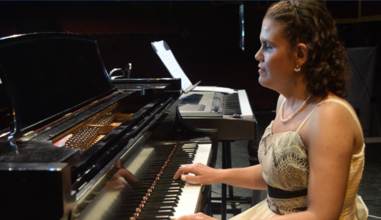 Una mujer con sordoceguera vestida con un traje de gala toca un piano de cola en el escenario; al fondo se ven unas partituras y otro teclado.