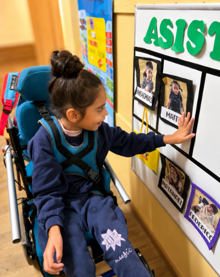 Niña sentada en una silla de ruedas frente a un panel de asistencia ubicado en la pared. Con una mano toca una de las imágenes del panel, que incluye fotografías y nombres escritos en imprenta mayúscula.