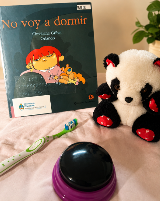 Libro infantil titulado No voy a dormir apoyado sobre una superficie, acompañado por un oso de peluche blanco y negro, un cepillo de dientes y un pulsador grande, como parte de una propuesta de lectura con objetos concretos.