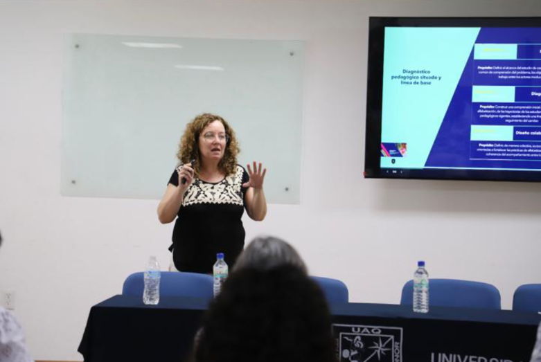 Screenshot 2026-06-29 134942 Mary Vázquez, una mujer con el pelo rizado, está de pie y gesticula mientras hace una presentación frente a una gran pantalla y una pizarra blanca. Tres personas están sentadas frente a ella en una mesa en la que hay botellas de agua. El escenario parece ser una aula o una sala de conferencias.