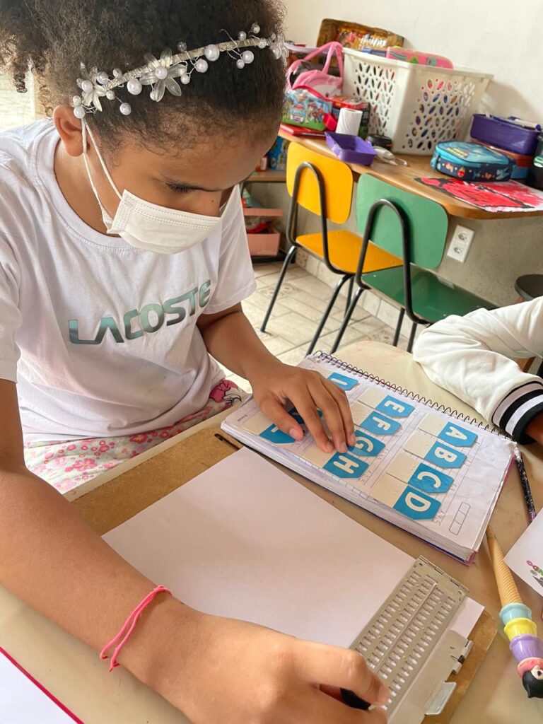 Ana Beatriz concentrada em suas tarefas escolares em uma mesa. Com a mão esquerda, ela toca letras em relevo em um caderno adaptado, que mostra as sequências "ABCD" e "EFGH" em blocos azuis. Com a mão direita, ela utiliza reglete metálica e punção presos a uma folha de papel em uma prancheta para escrever.