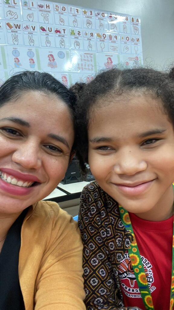 Uma selfie mostra Ana Paula e Ana Beatriz sorrindo lado a lado. À esquerda, Ana Paula tem cabelos escuros presos para trás e um sorriso largo. Ela está vestindo uma blusa preta por baixo de um cardigã na cor  mostarda. À direita, a menina, Ana Beatriz, tem cabelos escuros e cacheados presos em dois coques altos e sorri suavemente com os lábios fechados. Ela está vestindo uma camiseta vermelha com estampa gráfica e um cordão com estampa de girassol ao redor do pescoço. Ao fundo, ligeiramente fora de foco, há uma parede branca de sala de aula com cartazes educativos ilustrando o alfabeto em Libras (Língua Brasileira de Sinais).