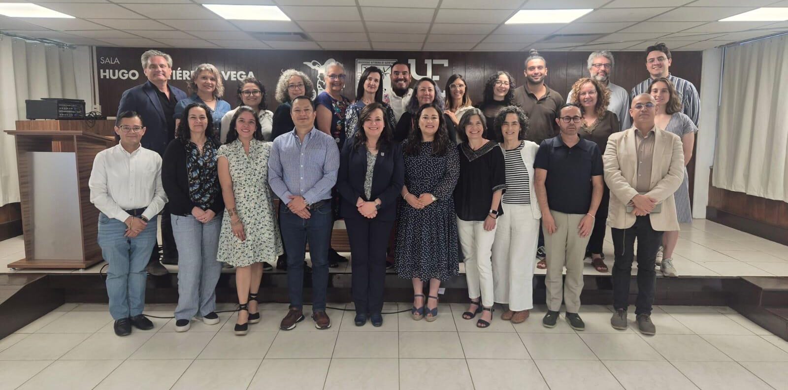 Un grupo de unas 25 personas, vestidas con ropa informal de oficina, posa sonriendo en una sala de conferencias con un atril a la izquierda y un cartel en la pared detrás de ellas.