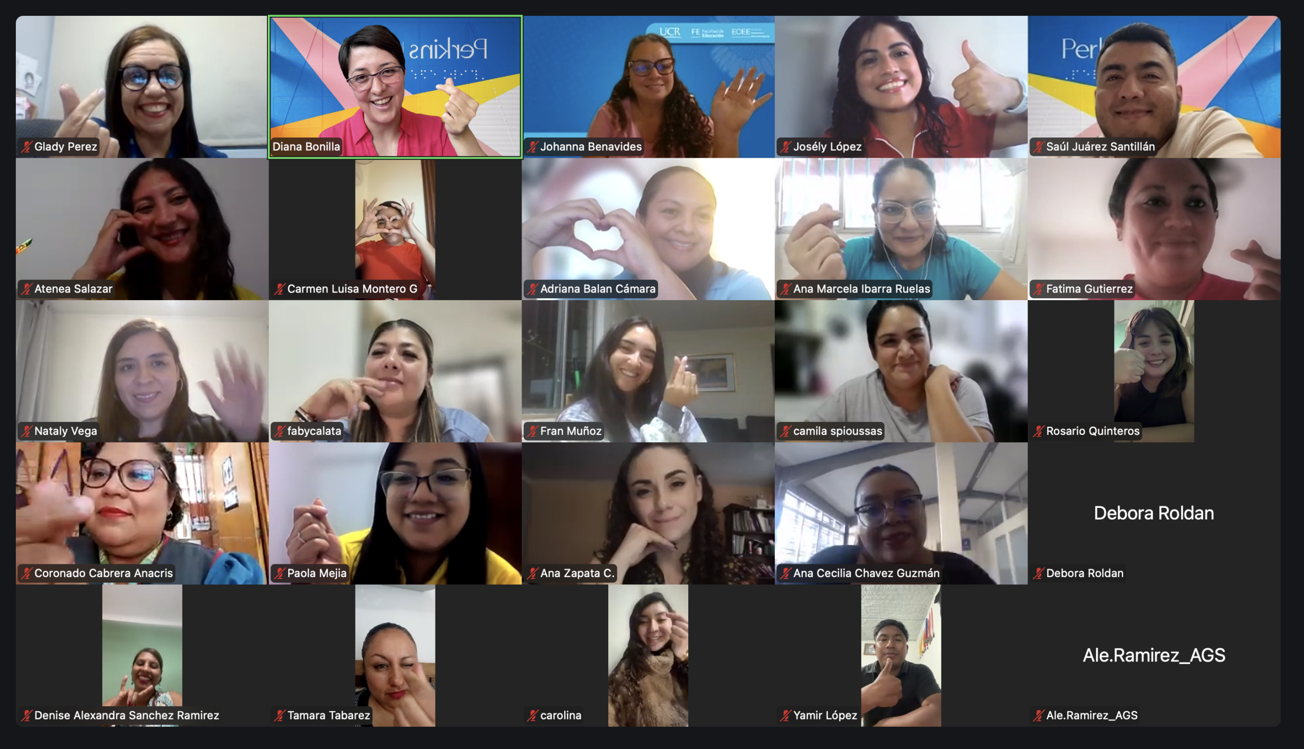 Una videollamada por Zoom con 25 participantes, la mayoría de los cuales sonríen, saludan con la mano o forman corazones con las manos. Los nombres aparecen debajo de cada imagen de vídeo. El fondo muestra entornos domésticos y de oficina.