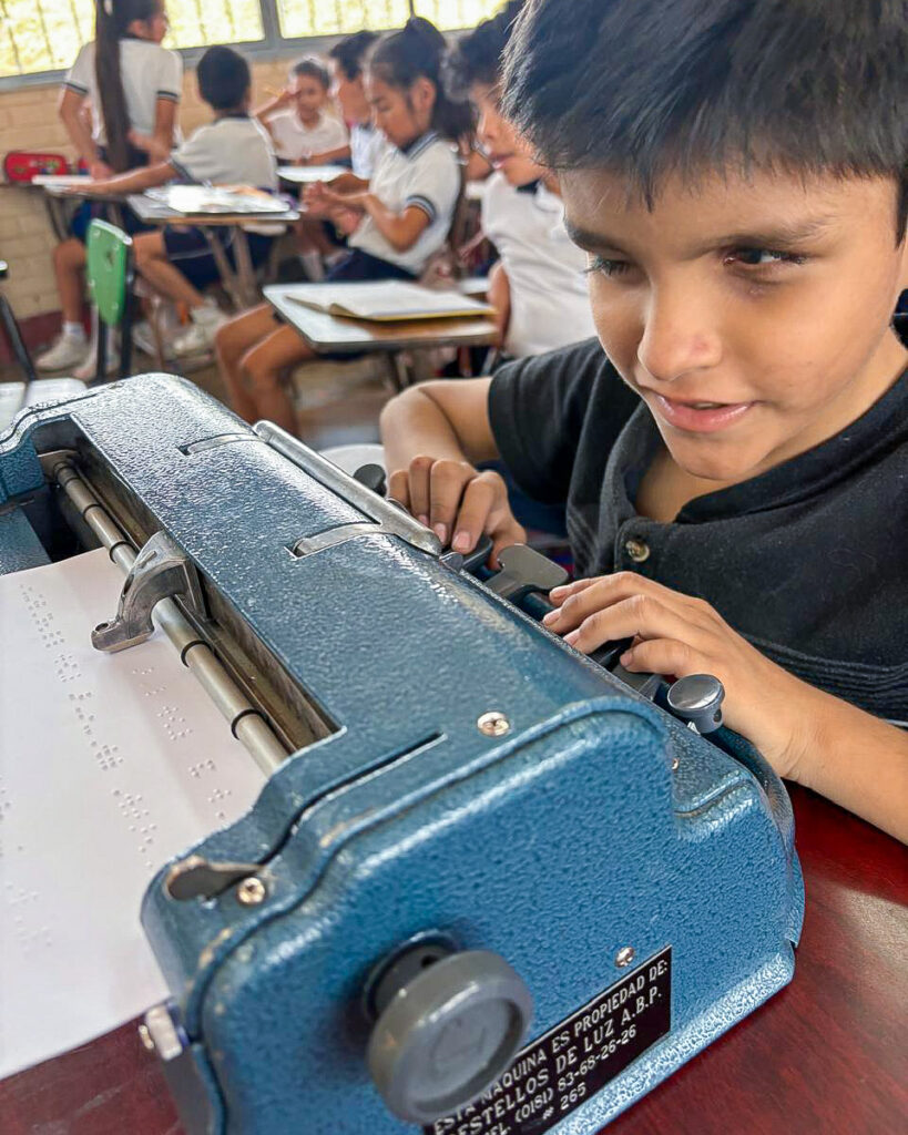 Luis utiliza una máquina Braille en un aula, mientras otros alumnos, sentados en sus pupitres, estudian al fondo.