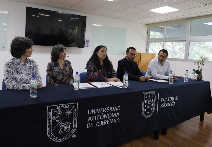 Cinco personas están sentadas alrededor de una mesa cubierta con un mantel azul oscuro en el que figuran los logotipos de la Universidad Autónoma de Querétaro y de la Facultad de Psicología; tienen delante documentos y botellas de agua, en una sala bien iluminada.