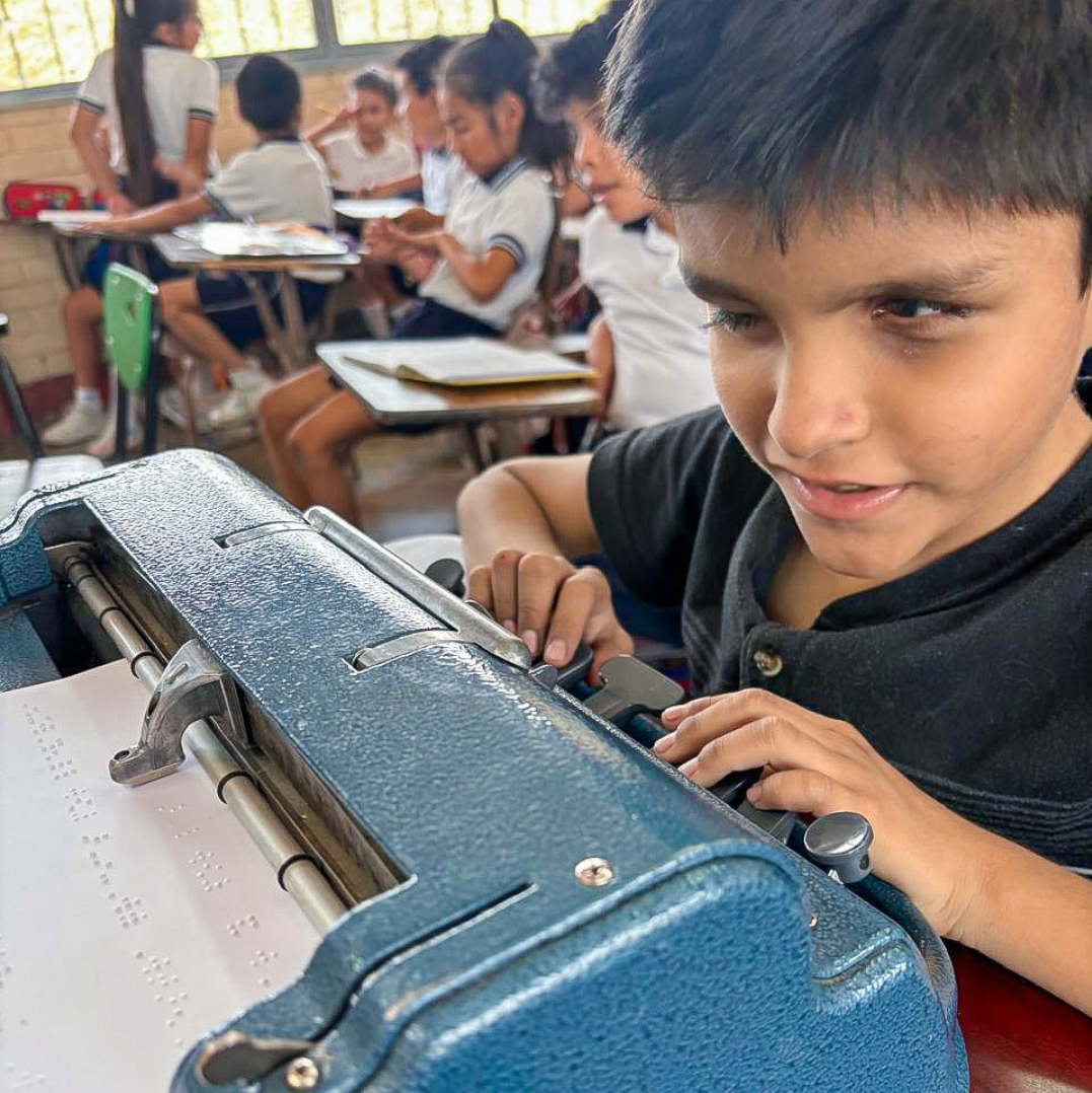 Luis utiliza una máquina Braille en un aula, mientras otros alumnos, sentados en sus pupitres, estudian al fondo.