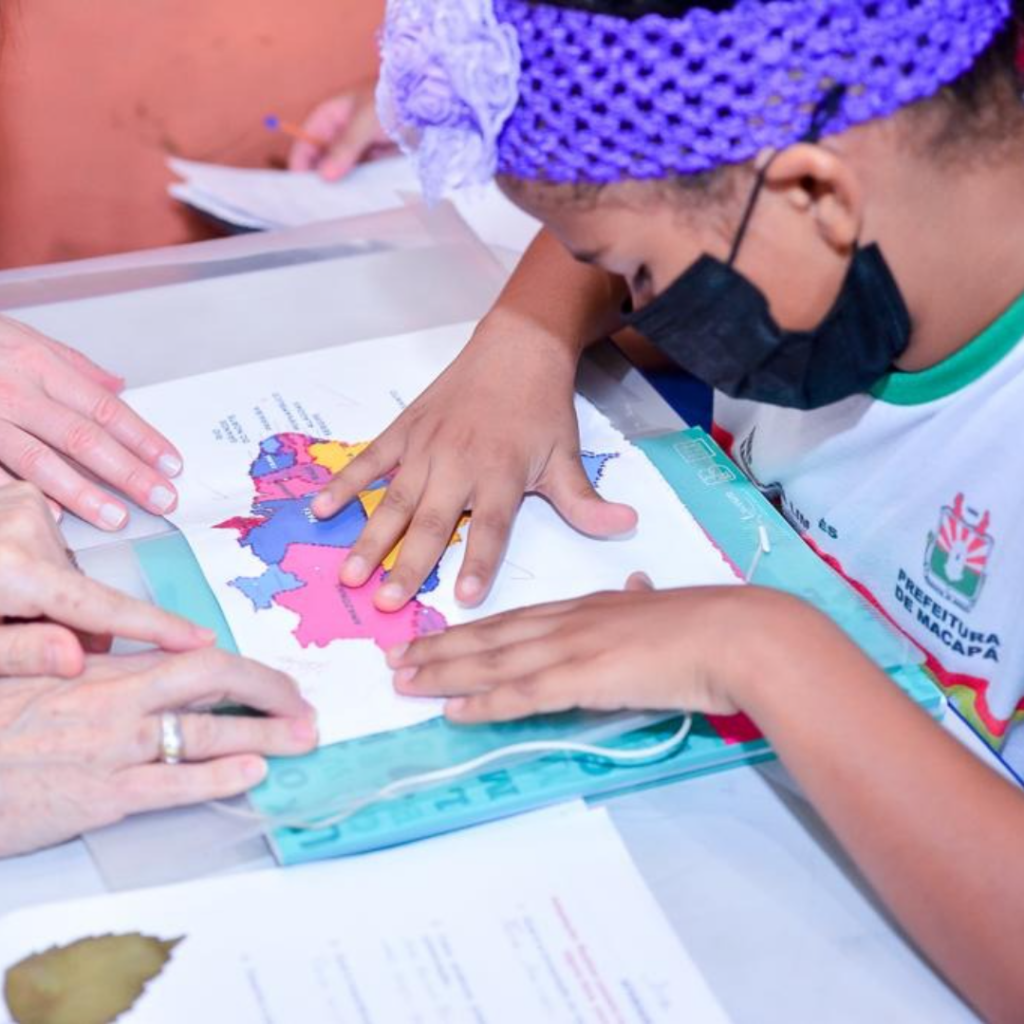 A foto mostra as mãos de uma estudante e de duas educadoras trabalhando juntas sobre uma mesa. À direita, a aluna usa uma faixa de crochê roxa no cabelo com uma flor, uma máscara cirúrgica preta e um uniforme escolar branco e verde. Suas mãos estão espalmadas sobre um mapa tátil adaptado e colorido do Brasil, explorando suas texturas e divisões regionais. À esquerda, as mãos das educadoras tocam o mesmo mapa com delicadeza, orientando a exploração tátil da aluna.