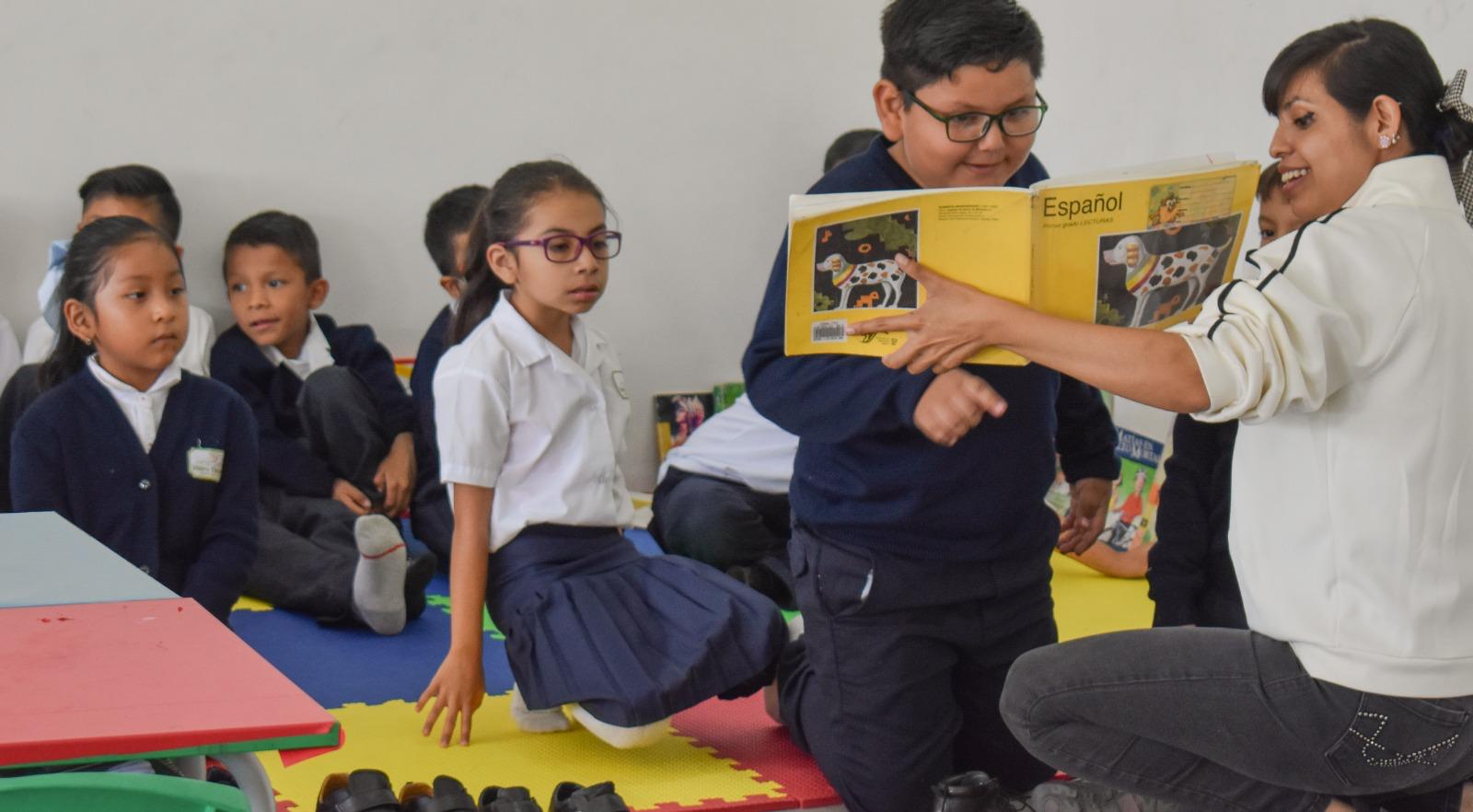 Una profesora lee un libro a un grupo de alumnos pequeños sentados sobre coloridas colchonetas de espuma. Los niños escuchan con atención y un niño se inclina hacia delante para mirar el libro.