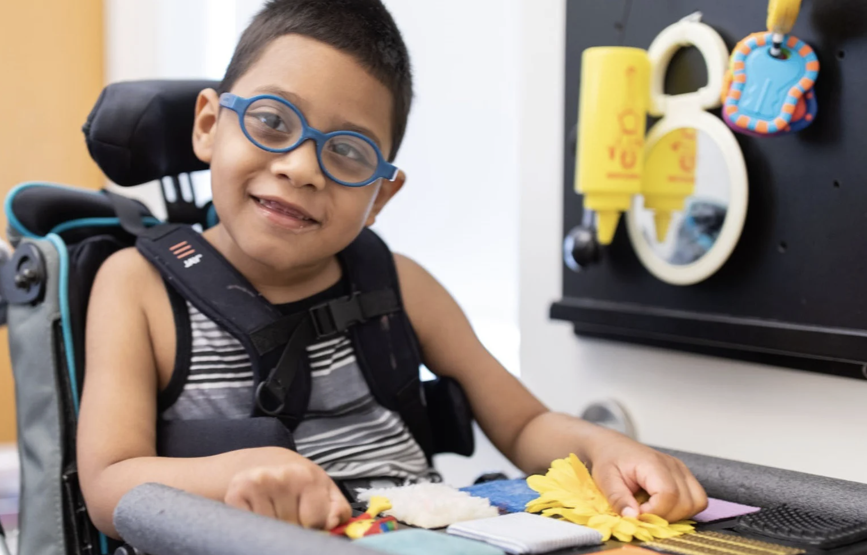 Un niño pequeño con gafas está sentado en una silla de ruedas, sonriendo y jugando con juguetes sensoriales texturizados en una bandeja. Un tablero de actividades sensoriales con espejos y objetos está montado en la pared junto a él.