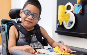 Un niño pequeño con gafas está sentado en una silla de ruedas, sonriendo y jugando con juguetes sensoriales texturizados en una bandeja. Un tablero de actividades sensoriales con espejos y objetos está montado en la pared junto a él.