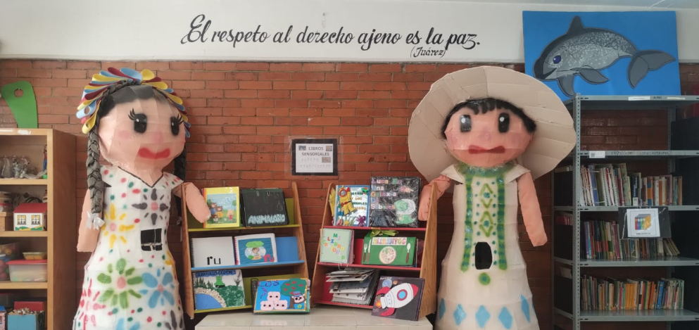 Screenshot 2026-09-08 163811 Dos grandes figuras de papel maché se alzan en una biblioteca llena de color. Detrás de ellas hay una cita escrita en la pared de ladrillo. En el fondo se ven estanterías, libros y un cuadro de un delfín.