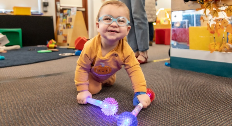 Un bebé sonriente con gafas gatea por una alfombra, sujetando dos juguetes luminosos y con púas, uno rojo y otro azul, en una colorida sala de juegos.