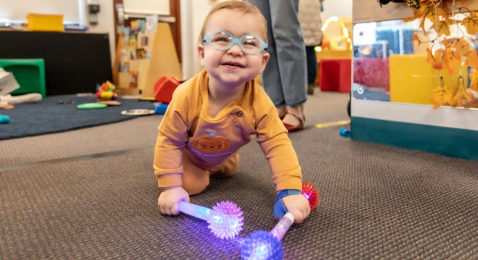 Un bebé sonriente con gafas gatea por una alfombra, sujetando dos juguetes luminosos y con púas, uno rojo y otro azul, en una colorida sala de juegos.