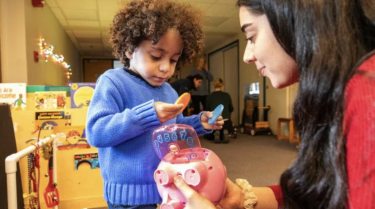 Un niño pequeño con un jersey azul introduce monedas en una hucha rosa que sostiene una mujer con un jersey rojo en un aula.